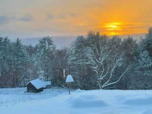 An orange sunset glows over a fresh wooded snow scene with a small snow-covered barn in the background and a snow-covered bird feeder in the foreground.