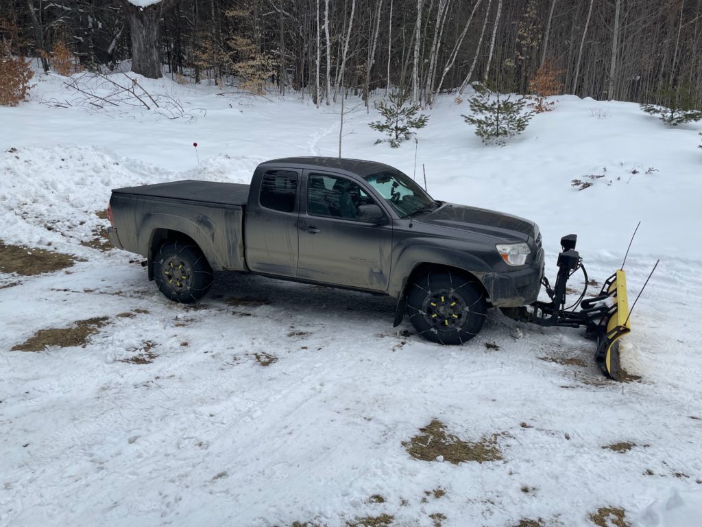 A dark grey 2014 Toyota Tacoma V6 pickup truck with chains on the tires and a yellow Fisher plow attached to the front.